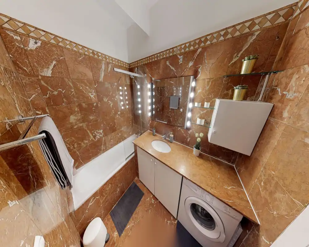 Baño compacto revestido en mármol con bañera, lavabo integrado en una encimera larga y lavadora debajo. Gran espejo con luces y estantes de cristal que aportan un estilo moderno y funcional.