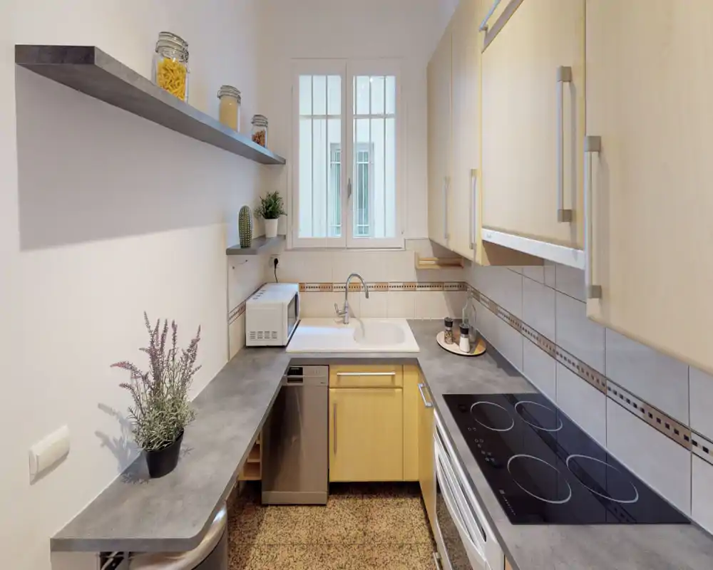 Cocina luminosa y bien equipada con placa eléctrica integrada, fregadero bajo la ventana, lavavajillas y armarios de madera clara — compacta y ordenada para el uso diario.