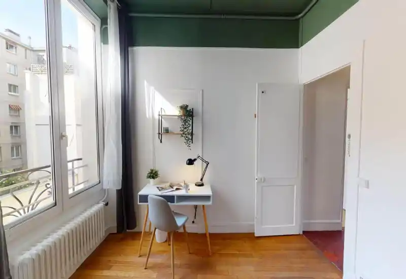 Pièce lumineuse avec un petit espace de travail près d'une grande fenêtre : bureau compact, chaise, lampe de bureau et plantes sur mur blanc, parquet et belle lumière naturelle, idéale pour étudier ou télétravailler.