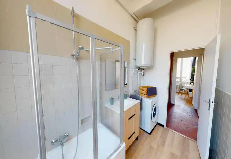 Clean, bright bathroom with a combined bathtub/shower, vanity with sink, washing machine and a water heater; wood-effect flooring and neutral tiles create a practical, well-lit space.