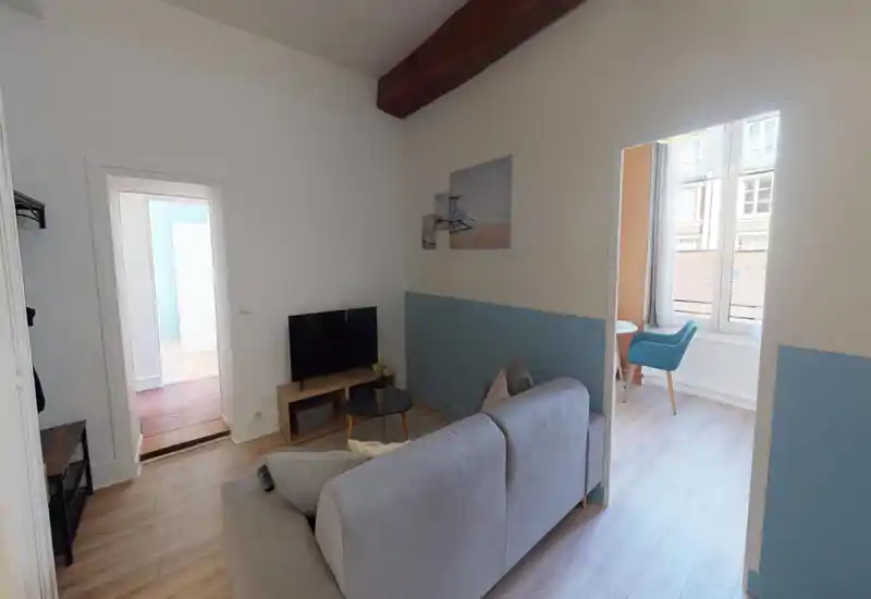 Salon lumineux et accueillant avec un canapé gris clair, une petite table basse et une télévision. La lumière naturelle de la grande fenêtre met en valeur le sol en bois et le mur accentué en bleu.