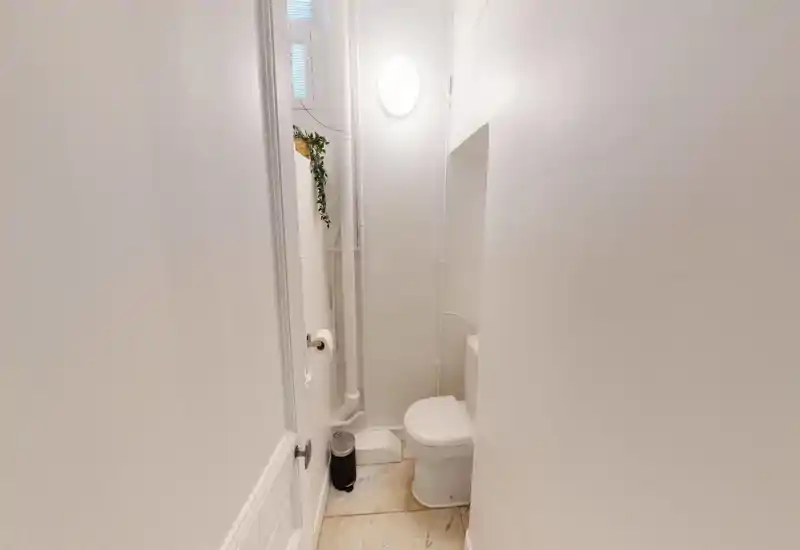 Compact, all-white guest toilet with visible plumbing, a wall light and a small trash bin; bright and clean minimal styling.