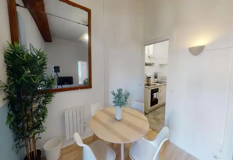 Bright, compact dining area with a round wooden table, four modern chairs and a decorative vase; opens to a small fitted kitchen — ideal for cozy meals.