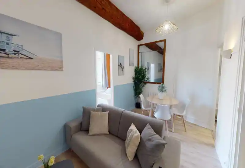 Bright, inviting living area with a comfortable grey sofa, round dining table and modern chairs. The room features tasteful coastal artwork, a large mirror, and exposed wooden beam giving a warm, stylish feel.