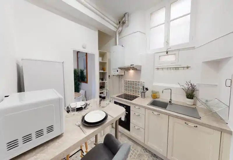 Bright compact kitchen with cream cabinets, built-in hob and sink. Patterned tile backsplash and a small breakfast counter set for two give a cozy, well-styled feel.