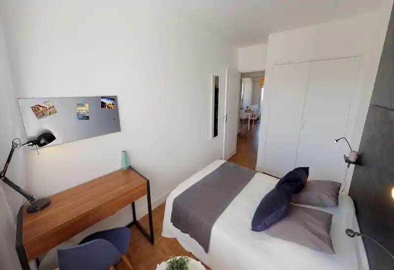 Bright, compact bedroom with a neatly made double bed, bedside reading lamps and a small wooden desk — ideal for a single occupant or couple. Clean white walls and warm parquet flooring give a calm, modern feel.