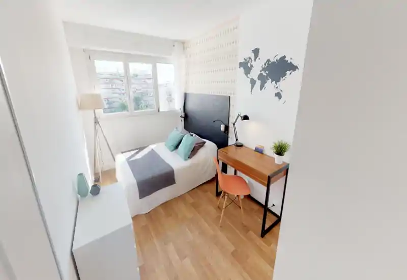 Sunny, compact bedroom with a neatly made double bed, large window providing abundant natural light, a small wooden desk and chair beside the bed, and tasteful minimal decor.