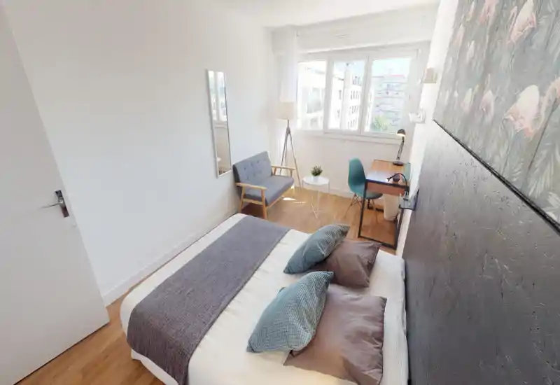 Chambre moderne et lumineuse avec lit double, coussins décoratifs et plaid texturé ; un petit coin salon et un bureau près de la fenêtre offrant une belle lumière naturelle et vue sur la ville.