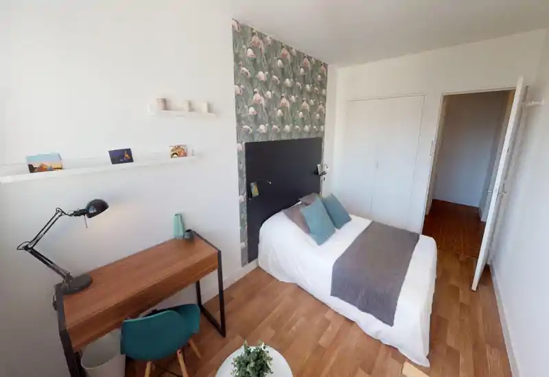 Chambre simple claire et bien décorée avec lit bien fait, papier peint flamants derrière la tête de lit, petit bureau en bois et chaise — idéale pour une location chaleureuse.