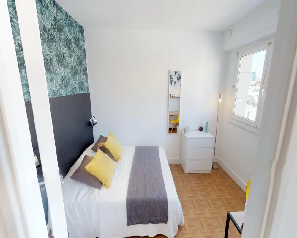 Camera da letto luminosa e arredata con un letto matrimoniale in lenzuola bianche, runner grigio e cuscini gialli. La stanza ha una parete testata decorata, pavimento in parquet, comò e luce naturale dalla finestra.