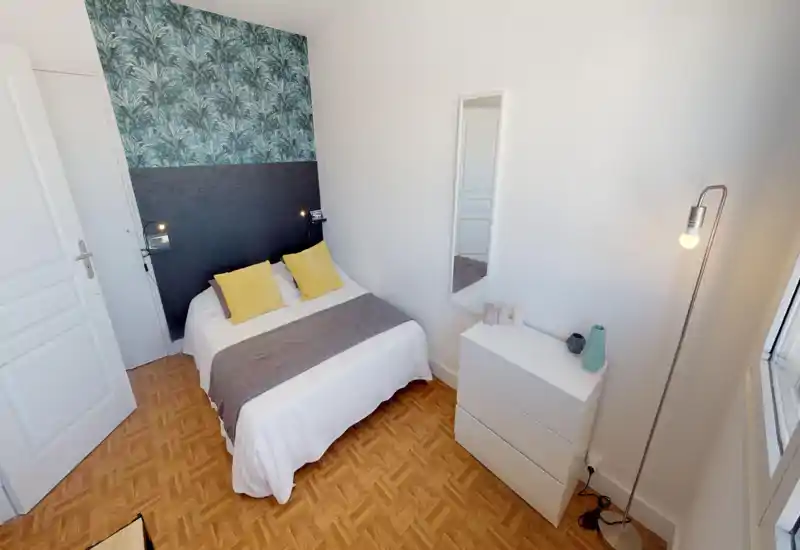 Bright, neatly staged double bedroom with patterned accent wall, bedside lights and cozy textiles. The bed is central and fully visible, with complementary cushions and a simple dresser under a tall mirror.