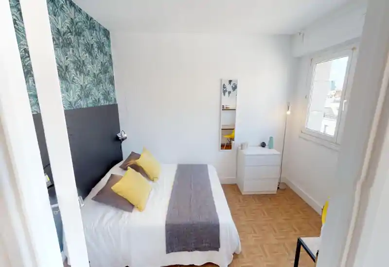 Bright, styled bedroom with a double bed dressed in white linen, gray runner and yellow accent pillows. The room features a patterned headboard wall, parquet floor, bedside chest and natural light from a window.