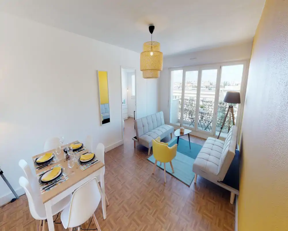 Bright, staged living room with two sofas, a small coffee table and a yellow accent chair facing large floor-to-ceiling windows that open onto a balcony; an adjacent dining table set for four completes the open-plan space.