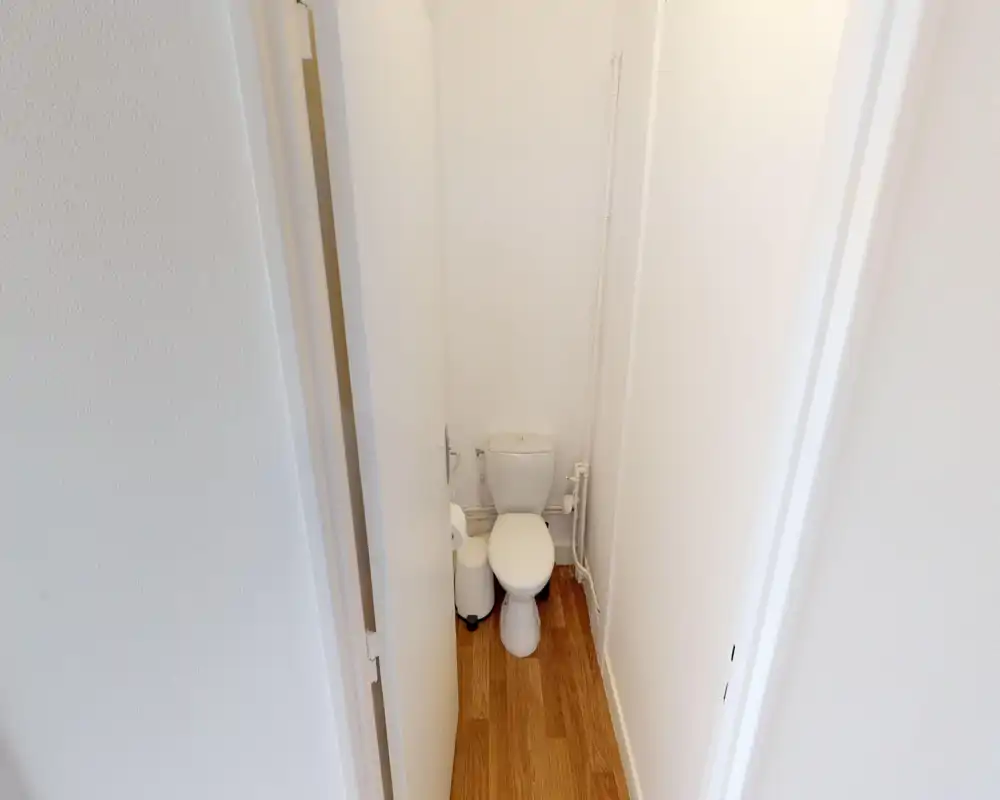 Compact bathroom with a standard toilet in a narrow, clean space featuring white walls and wood-effect flooring.