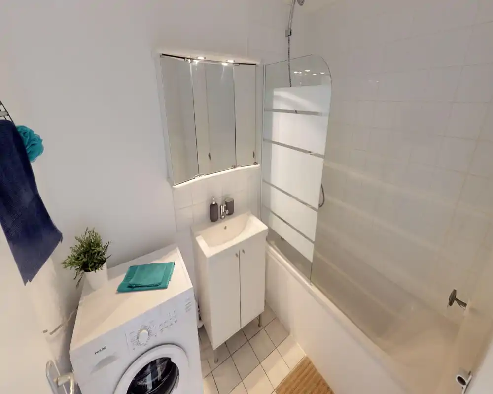 Compact, clean bathroom with a bathtub and glass shower screen, small sink cabinet and a washing machine tucked into the corner. Neutral tiles and white fixtures create a bright, tidy space suitable for rental listings.