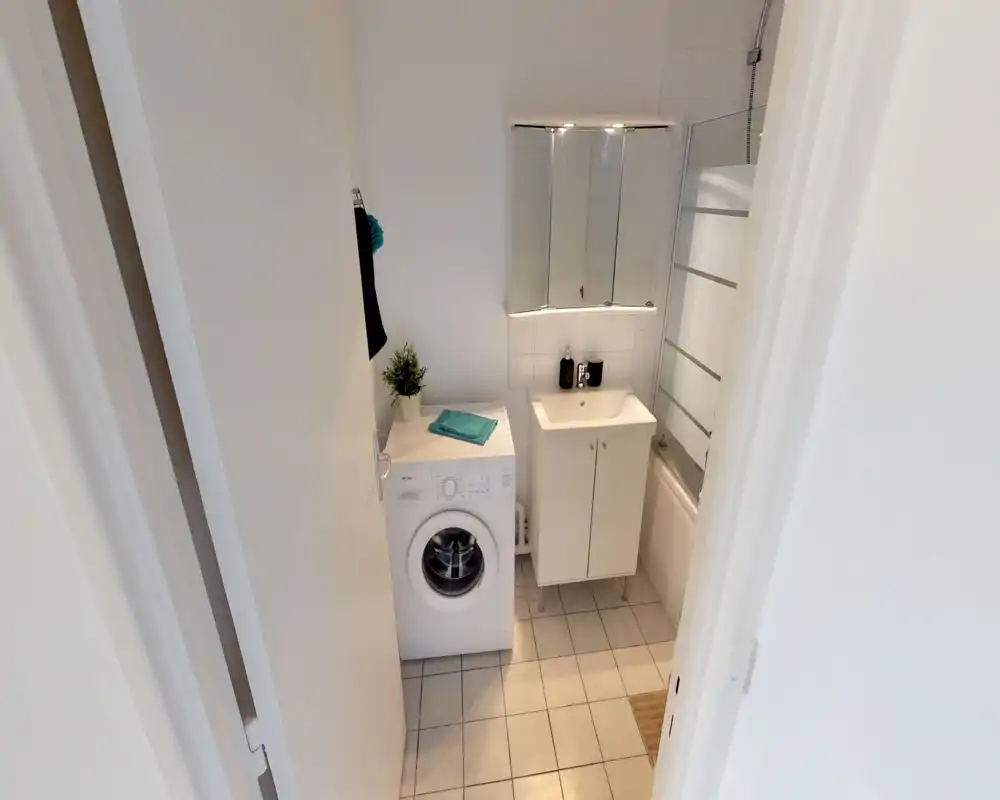 Compact, well-lit bathroom with a sink cabinet, mirrored medicine cabinet, bathtub with glass screen and a washing machine — neat tiled floor and bright white finishes.