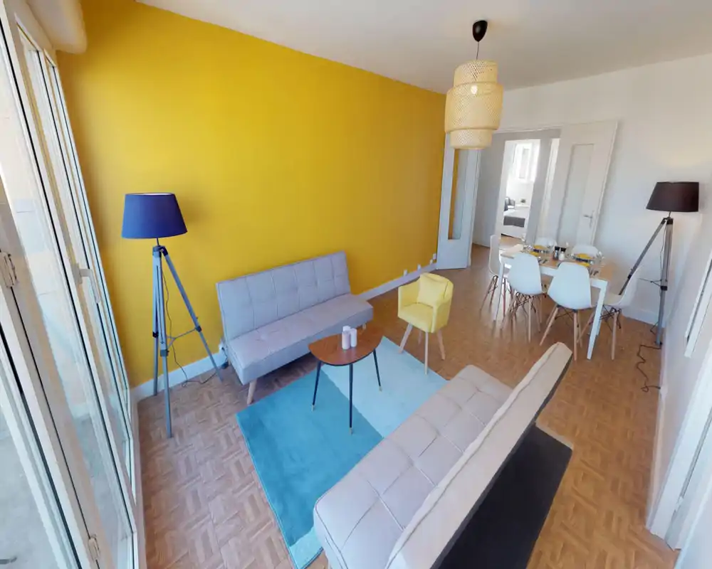 Spacious, stylish living room with a bold yellow accent wall, two modern sofas, a blue rug and a dining area in the background — bright natural light floods the space.