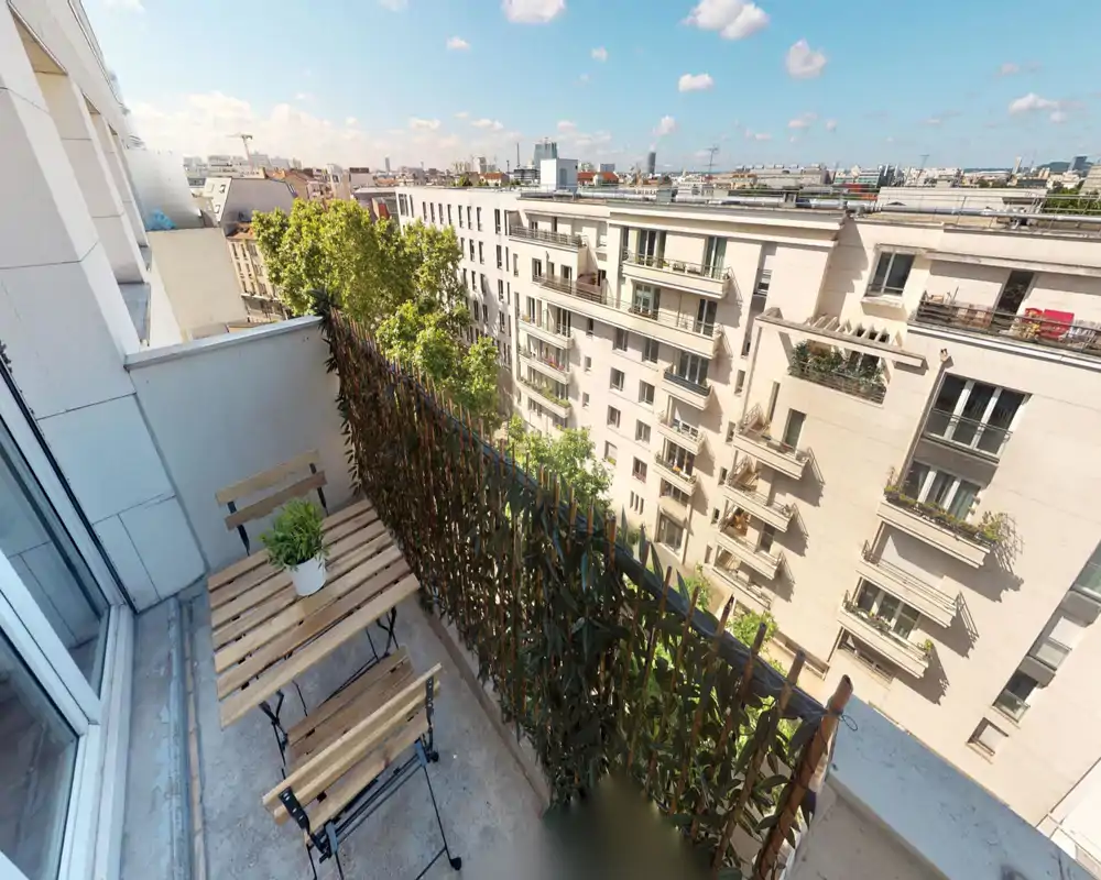 Sunny narrow balcony with a small wooden bistro table and two folding chairs, potted plant, privacy screen and a city skyline view — ideal for morning coffee.