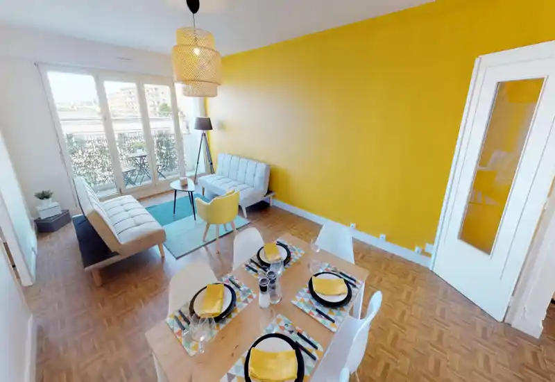 Séjour lumineux et bien mis en scène avec un mur d'accent jaune vif, deux canapés, une petite table basse et un coin repas au premier plan. De grandes baies vitrées s'ouvrent sur un balcon et inondent l'espace de lumière naturelle.