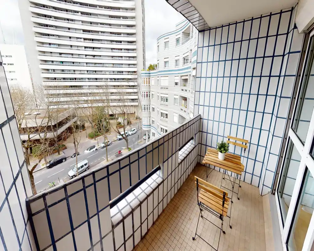 Balcone piastrellato con un piccolo tavolo in legno e due sedie pieghevoli, affacciato su una strada alberata e palazzi vicini — perfetto per il caffè mattutino.