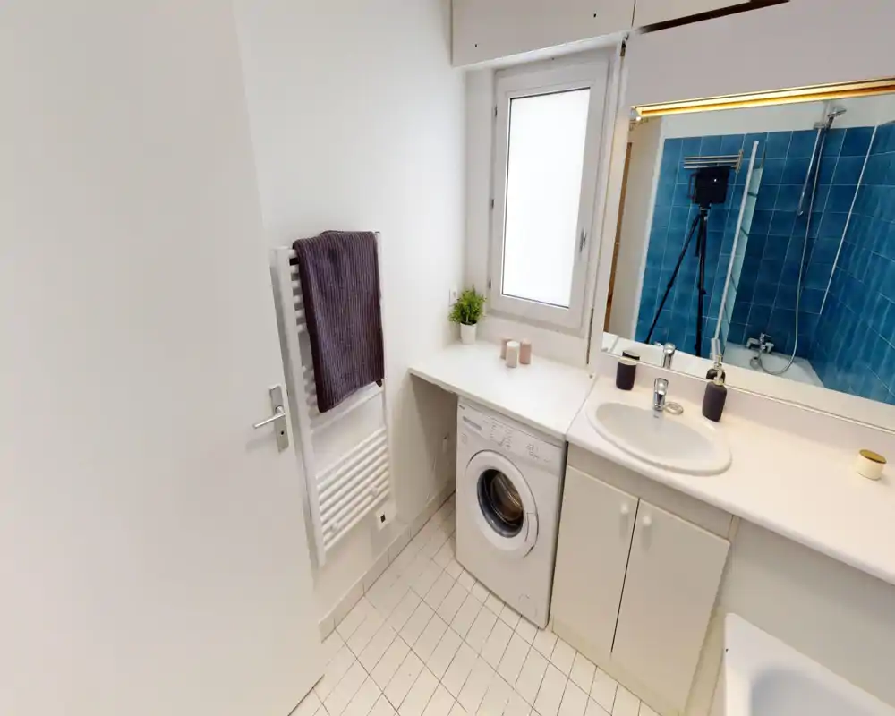 Bagno moderno e pulito con lavabo, piano di appoggio, lavatrice incassata e vasca con piastrelle blu visibili nello specchio. Spazio funzionale e luminoso con sanitari bianchi e pavimento piastrellato.