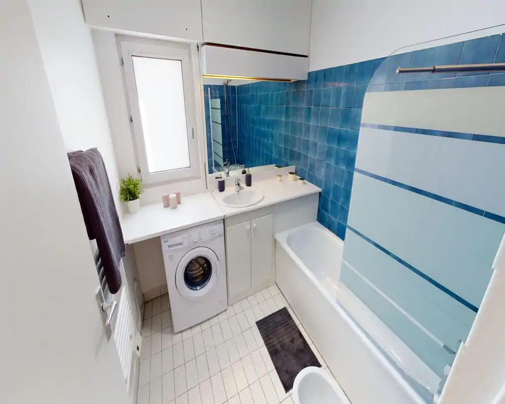 Bagno luminoso e ordinato con vasca, parete doccia, lavabo e lavatrice sotto il piano; parete piastrellata blu e finestra satinata per privacy e luce naturale.