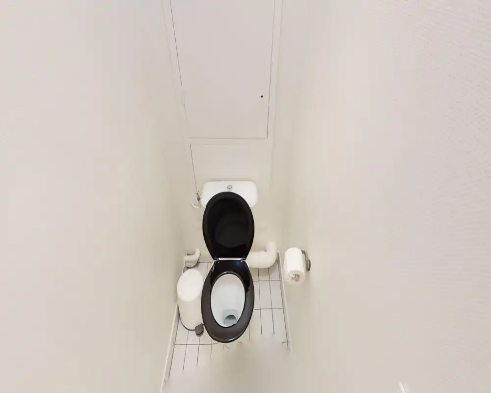 Bagno toilette compatto e pulito con wc a seduta nera, portarotolo a parete e piccolo cestino; pareti bianche e pavimento piastrellato donano un aspetto minimale e ordinato.