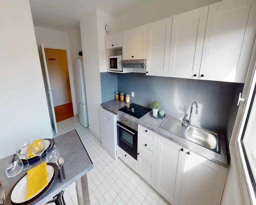 Cucina compatta e luminosa con mobili bianchi, piano cottura elettrico, forno incassato e lavabo in acciaio. Piccolo tavolo da colazione per due e barattoli decorativi rendono l’ambiente ordinato e accogliente.