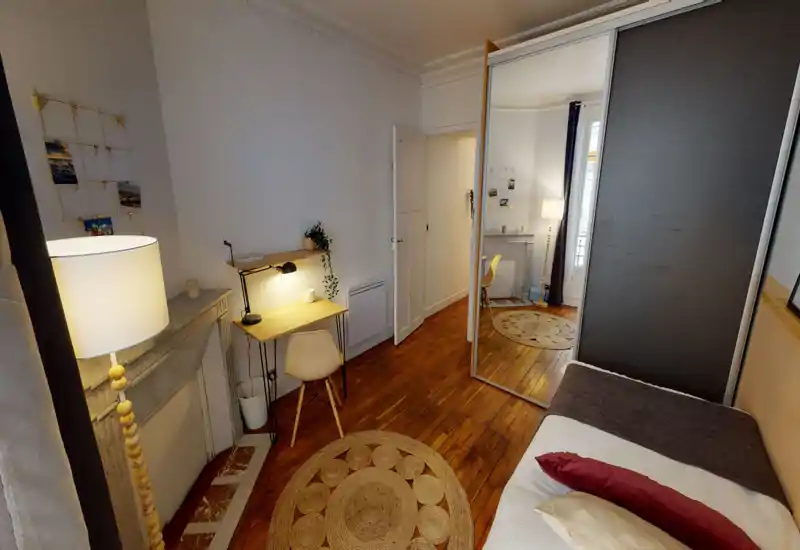 Camera da letto luminosa e accogliente con pavimenti in legno, un letto singolo con cuscini, una lampada da terra e una scrivania; l'armadio con specchio dona profondità.