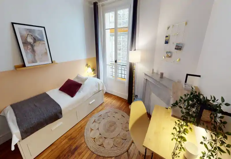 Camera da letto singola luminosa e ben arredata con letto confortevole, pavimento in legno, tappeto rotondo intrecciato e scrivania vicino alla finestra. Tessuti morbidi e illuminazione stratificata rendono l’ambiente accogliente e ideale per soggiorni brevi o lunghi.