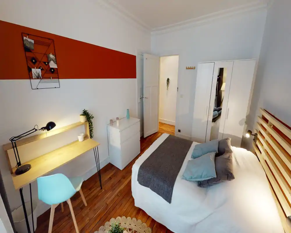 Chambre lumineuse et bien décorée avec un lit double, coussins superposés et une tête de lit en lattes de bois. La pièce comprend un bureau, une armoire blanche avec miroir, une commode et un parquet chaleureux — idéale pour des annonces mettant en avant confort et praticité.