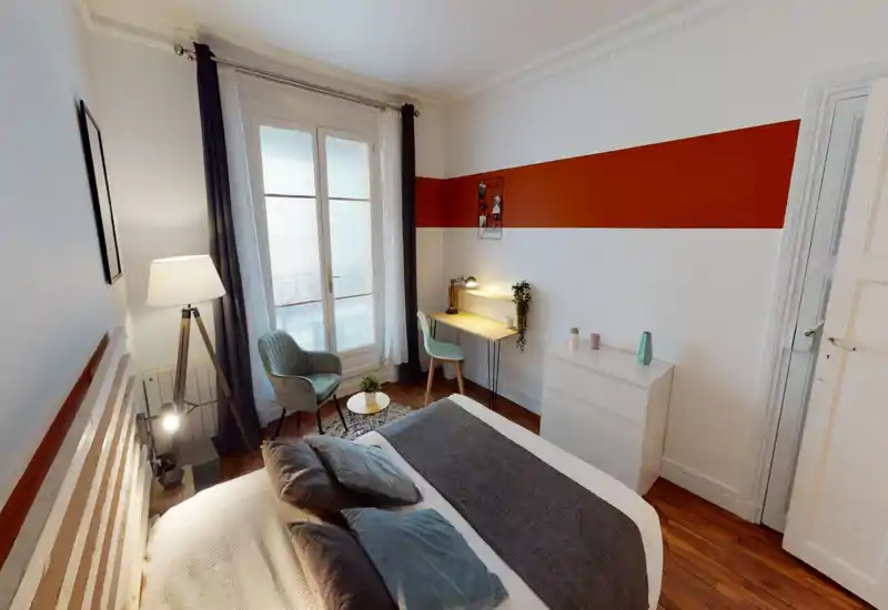 Bright, stylish bedroom with a large bed in the foreground, soft pillows, a bedside lamp, a seating corner and a small desk by the window — ideal for relaxing or remote work.