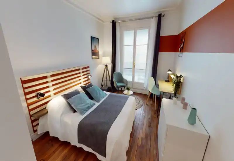 Bright, stylish bedroom with a large double bed, decorative cushions, wooden headboard and warm hardwood floors. A window/door with sheer curtains and a small seating area add natural light and a cozy atmosphere.