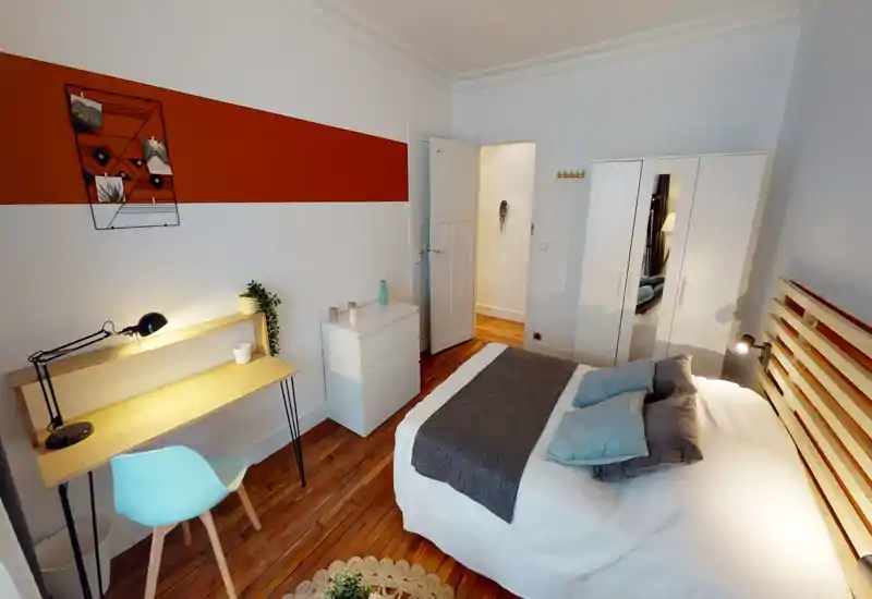 Bright, well-styled bedroom with a double bed, layered pillows and a wooden slatted headboard. The room features a desk workspace, white wardrobe with mirrored panel, chest of drawers and warm hardwood floors — ideal for rental listings highlighting comfort and functionality.