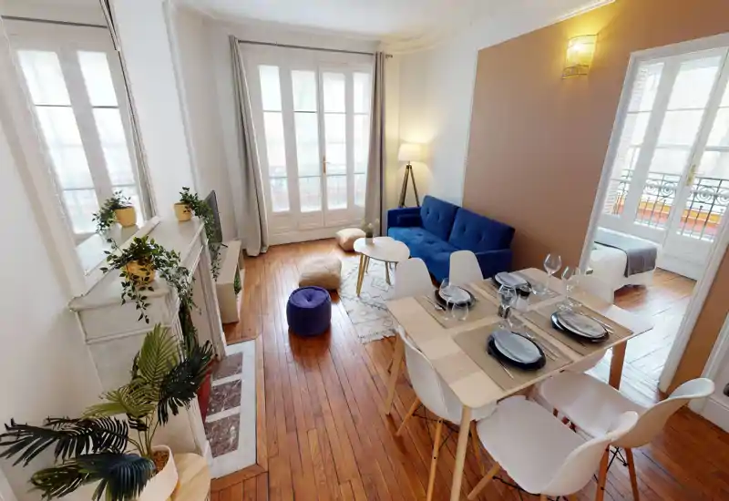 Bright, stylish living area with a blue sofa, coffee table and dining setup; large French windows bring abundant natural light and the wooden floors add warmth.