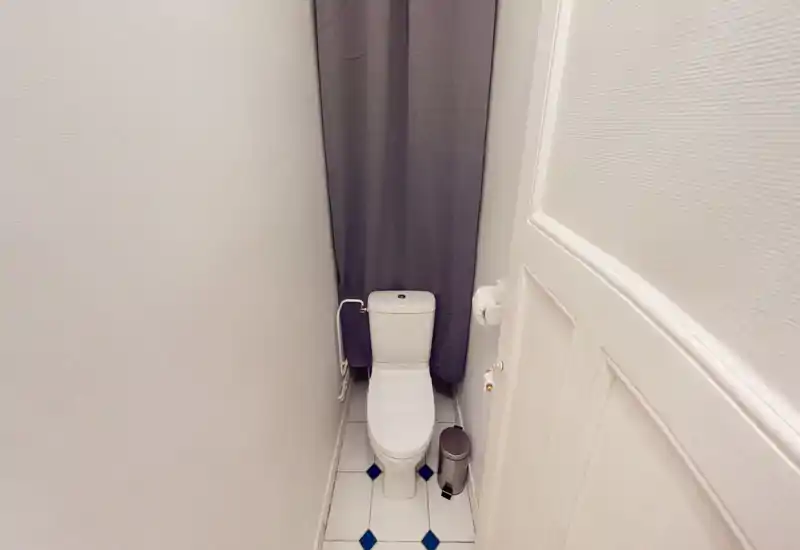 Compact, clean toilet room with a white ceramic WC, small trash bin and neutral tiled floor; fitted with a dark curtain at the back.
