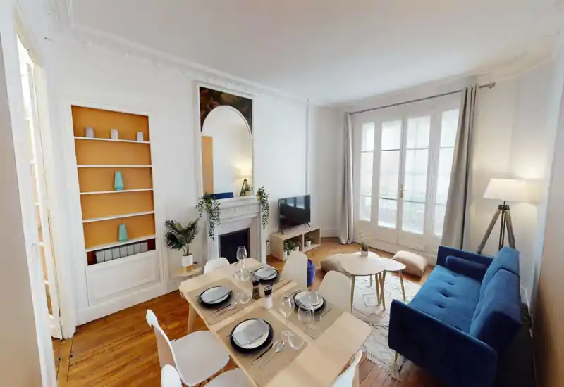 Spacious, well-lit living and dining area with a cozy blue sofa, wooden dining table set for four, decorative rug and large French windows that flood the room with light.