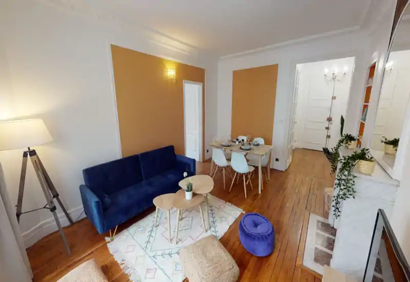 Bright, stylish living room with a blue velvet sofa, nested wooden coffee tables, patterned rug and a dining table set in the background — welcoming and well-styled for rental photos.