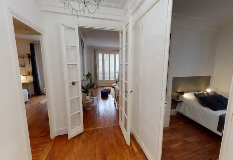 Angolo camera luminoso e elegante visibile attraverso porte a vetri, con letto ben fatto, lampade da comodino e pavimenti in legno che conducono a una zona giorno soleggiata.