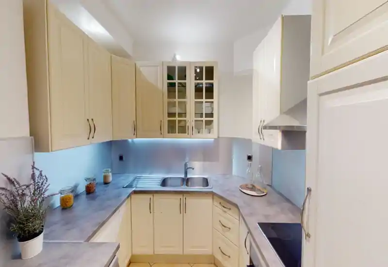 Well-lit modern kitchen with cream cabinets, integrated sink, glass-front upper cupboards and tidy countertops with decorative jars and a potted plant.