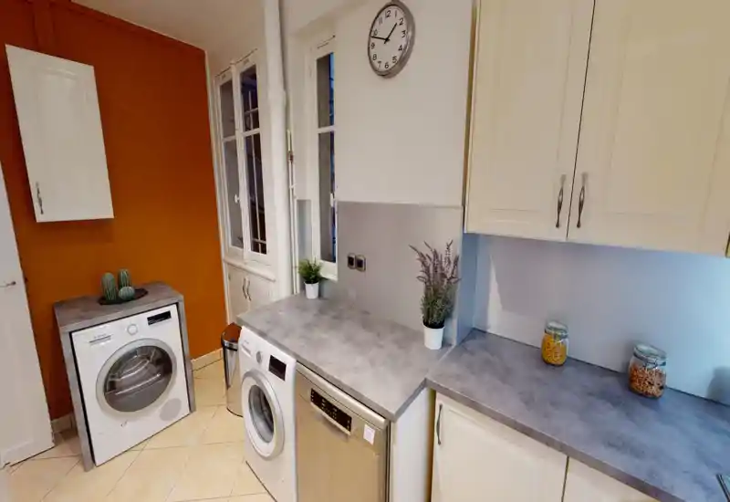 Bright, tidy kitchen-laundry corner with integrated appliances. Countertops with decorative jars and plants create a neat, welcoming utility space.