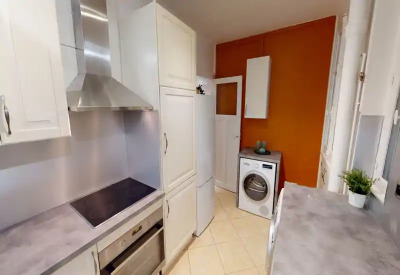 Well-equipped compact kitchen with modern stovetop, oven and hood, a refrigerator and a washer; warm orange accent wall and light countertops create a cozy, functional space.