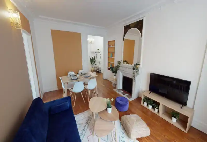 Bright, stylish living room with a deep-blue sofa, nested wooden coffee tables, a TV unit and a dining table in the background. Warm wooden floors, decorative plants and a fireplace create a cozy, well-staged space.