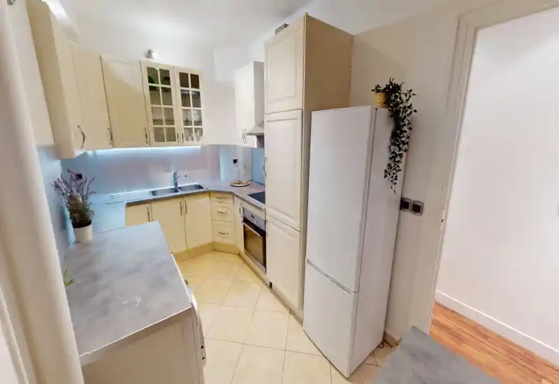 Compact, well-equipped kitchen with cream cabinetry, built-in oven, induction hob and full-height fridge; under-cabinet lighting and decorative plants create a tidy, bright space.