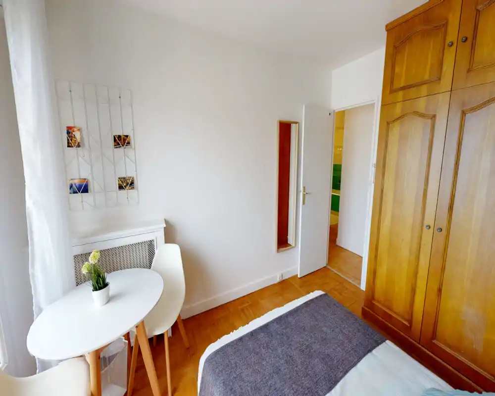 Bright, neatly staged bedroom with a visible bed corner, wooden wardrobe and a small round table by the window offering natural light.