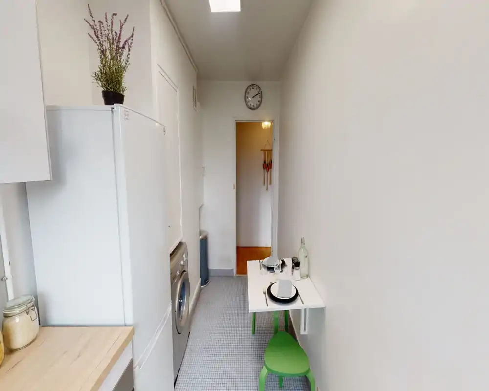 Narrow galley-style kitchen with white cabinets, a fridge, washing machine and a small fold-down table set for two. Clean minimalist styling with green stool accents and good natural/ambient light.