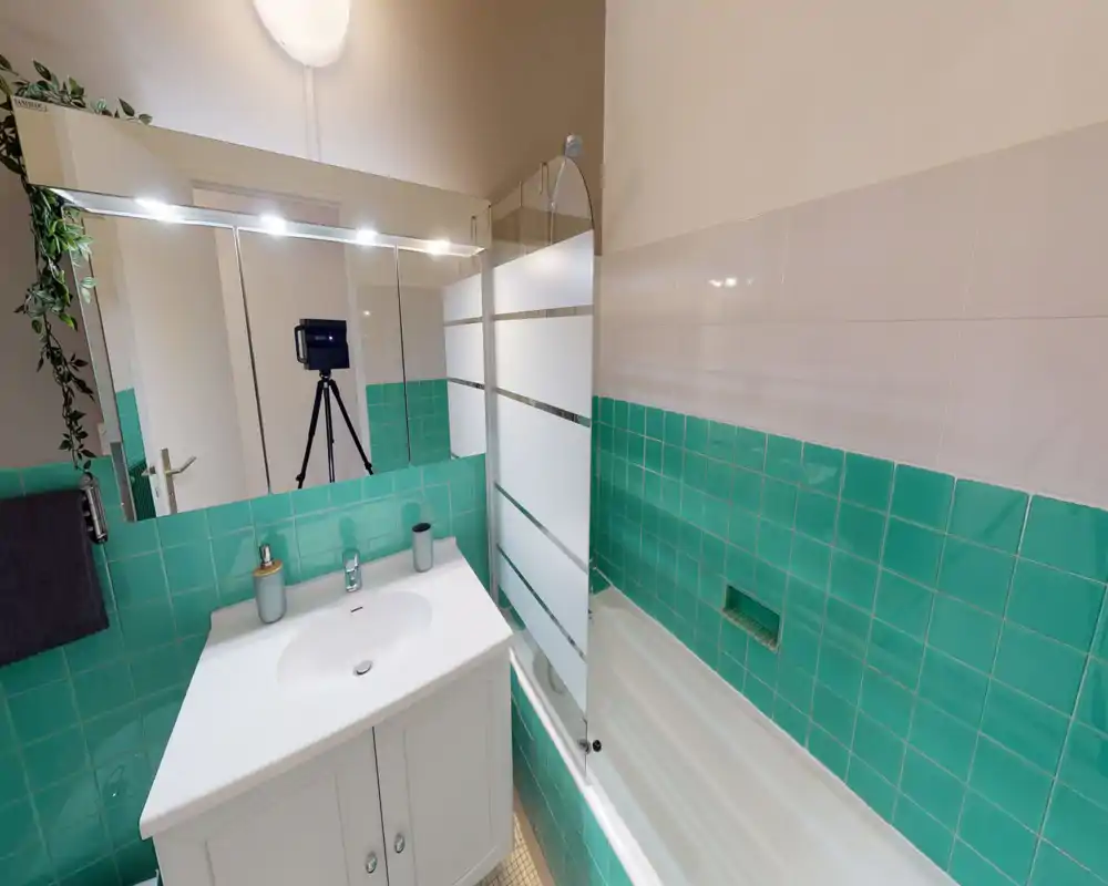 Clean, bright bathroom with turquoise wall tiles, a white vanity sink, and a bathtub with a glass shower screen. Large mirror and modern fixtures create a fresh, well-maintained feel.