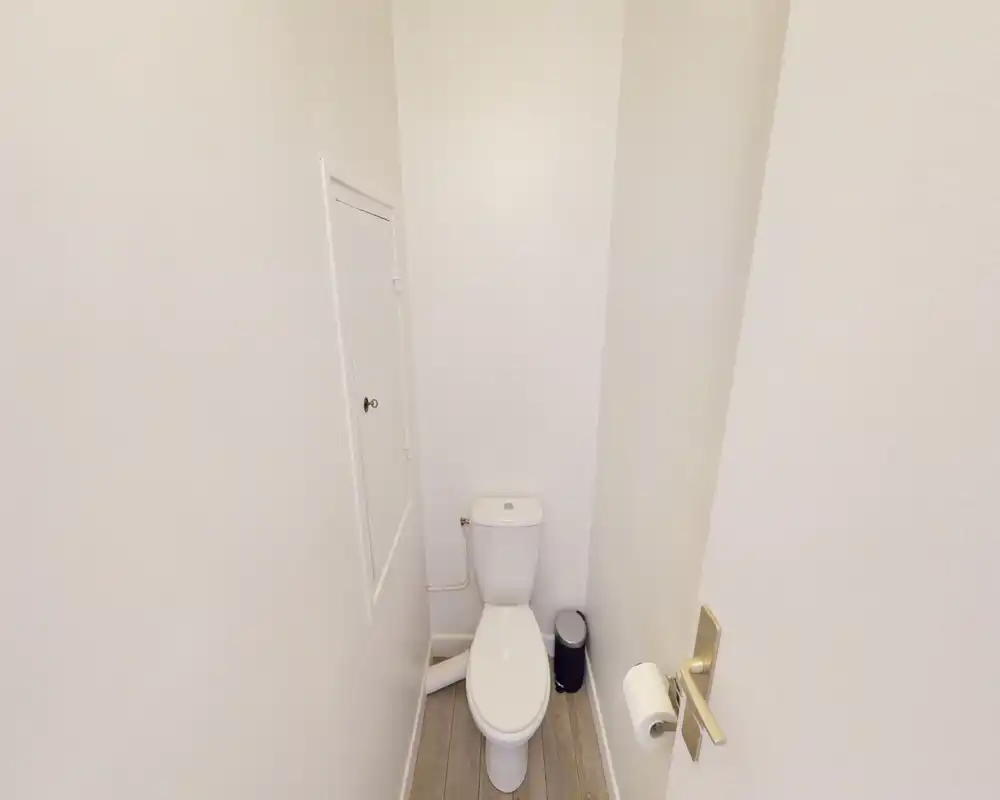 Compact half-bath with a single white toilet, small waste bin and wall-mounted toilet paper holder. Neutral white walls and light wood-look floor create a clean, minimal look.