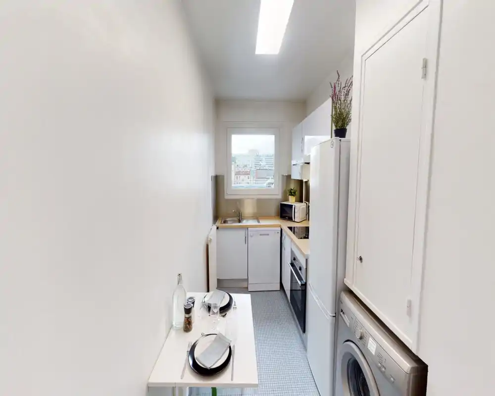 Bright, compact galley kitchen with integrated appliances, a small dining surface set for two, and a washing machine — ideal for a city apartment. Clean, modern finishes and a window providing natural light.