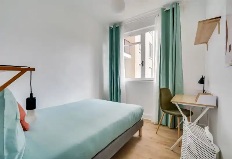 Bright, modern single bedroom with a neatly made bed in soft teal linens, a small desk by the window and calming mint curtains — ideal for a comfortable stay.